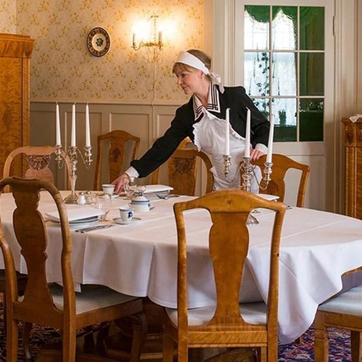Stuepige i uniform dækker bord med hvid dug, keramisk service og sølvlysestage i elegant spisestue.