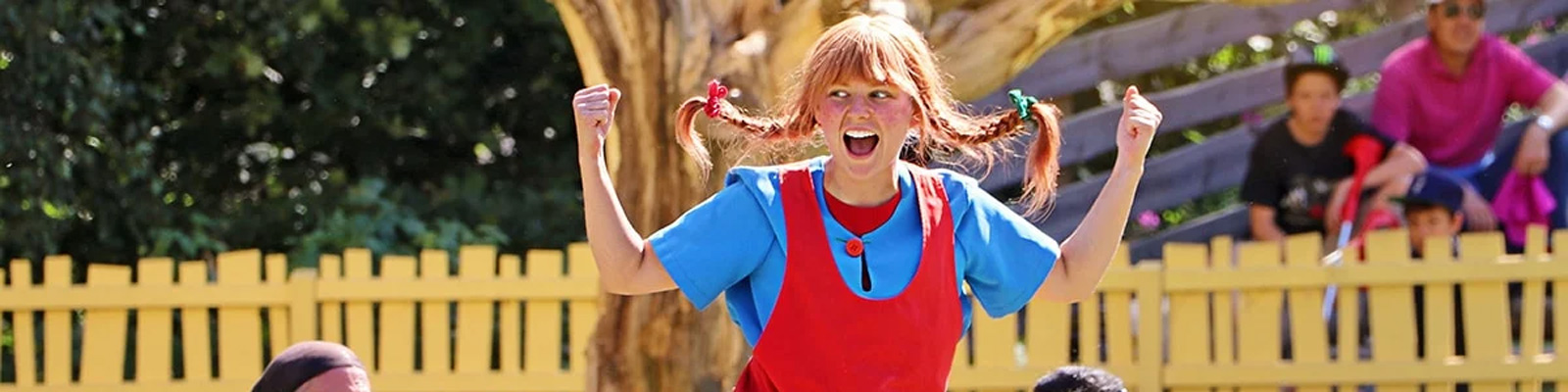 Pige klædt som Pippi Langstrømpe i farverig udklædning jubler udendørs i solskin.