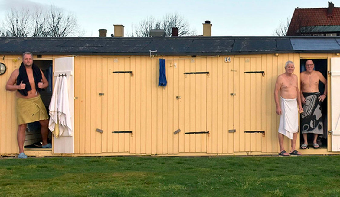 Mænd i badehåndklæder står foran en traditionel badstue ved stranden på en kølig dag i Danmark.