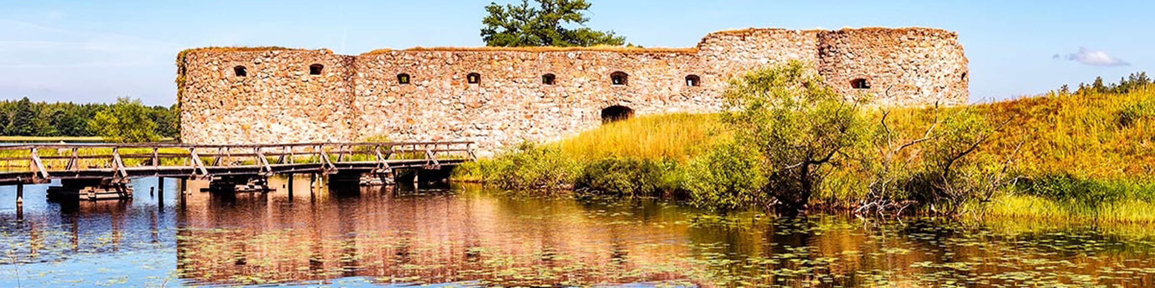 Ruiner af middelalderborg beliggende ved en stille sø i solrigt landskab med blå himmel.
