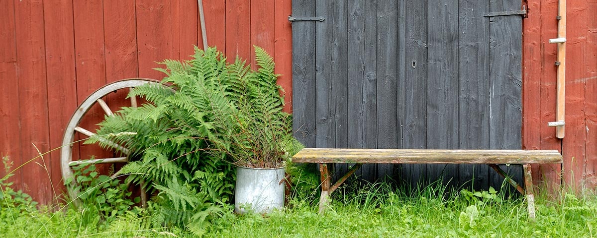 Foran en rød træbeklædning står en bænk, et træhjul og planter.