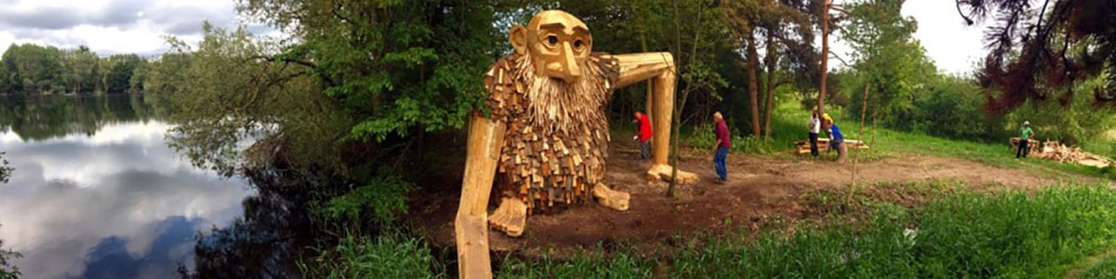 Jätteträfigur i park vid sjö omgiven av träd, skulpterad av trä och observerad av tre personer.