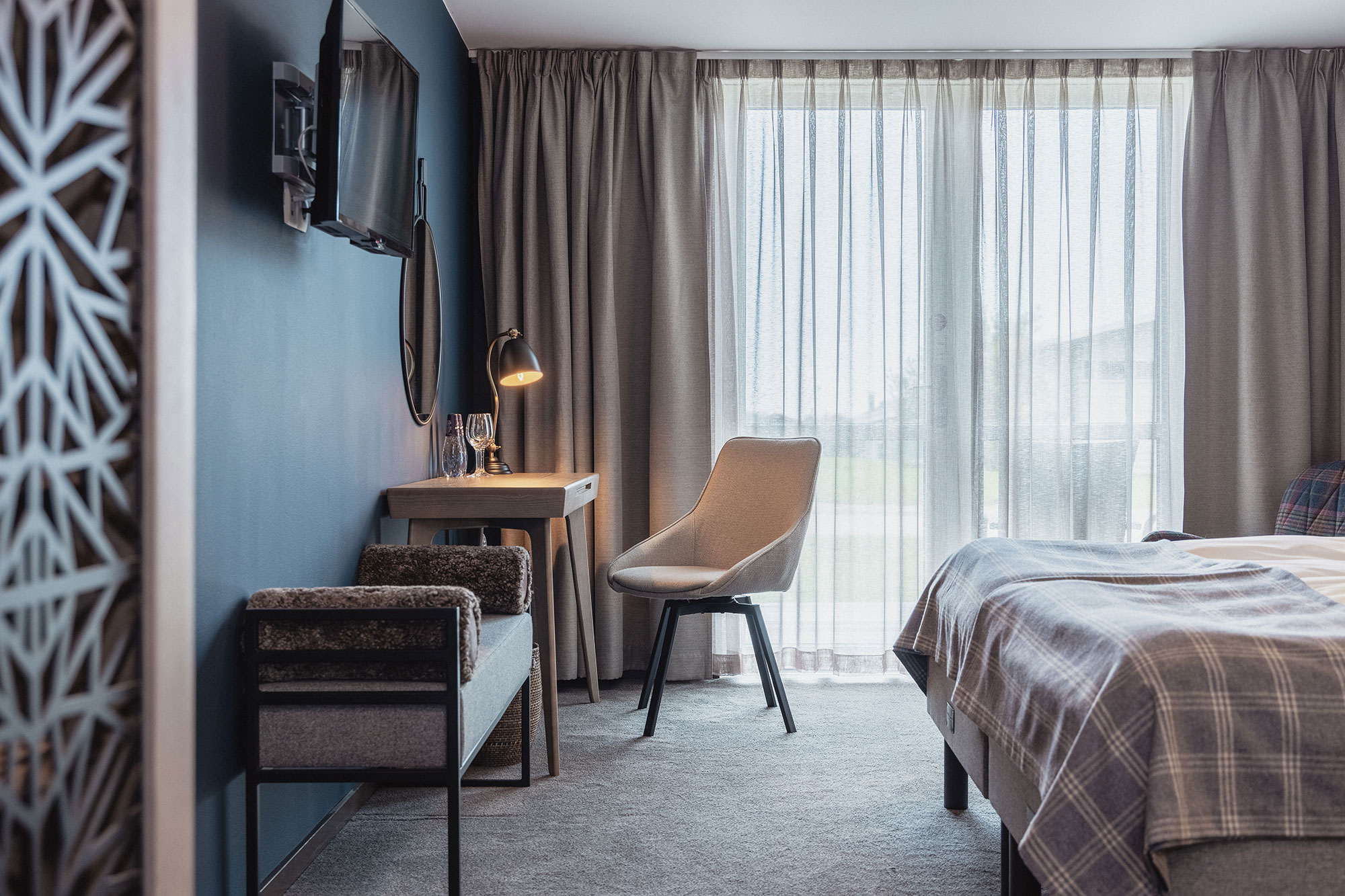 Moderne hotelværelse med skrivebord, stol og seng ved siden af store vinduer med lysindfald.