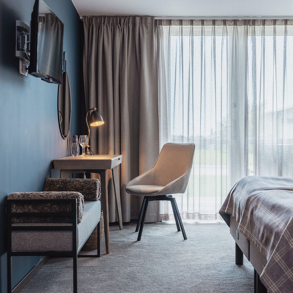 Moderne hotelværelse med skrivebord, stol og seng ved siden af store vinduer med lysindfald.