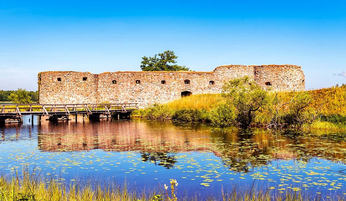 Gammel stenslot ved vand med åkander og en træbro, prydet af klar blå himmel i et naturskønt landskab.