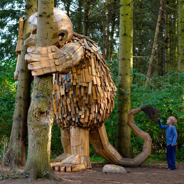 Träskulpturen Tilde av Kph Jattetrollen omfamnar ett träd medan ett barn tittar på i skogsmiljö.