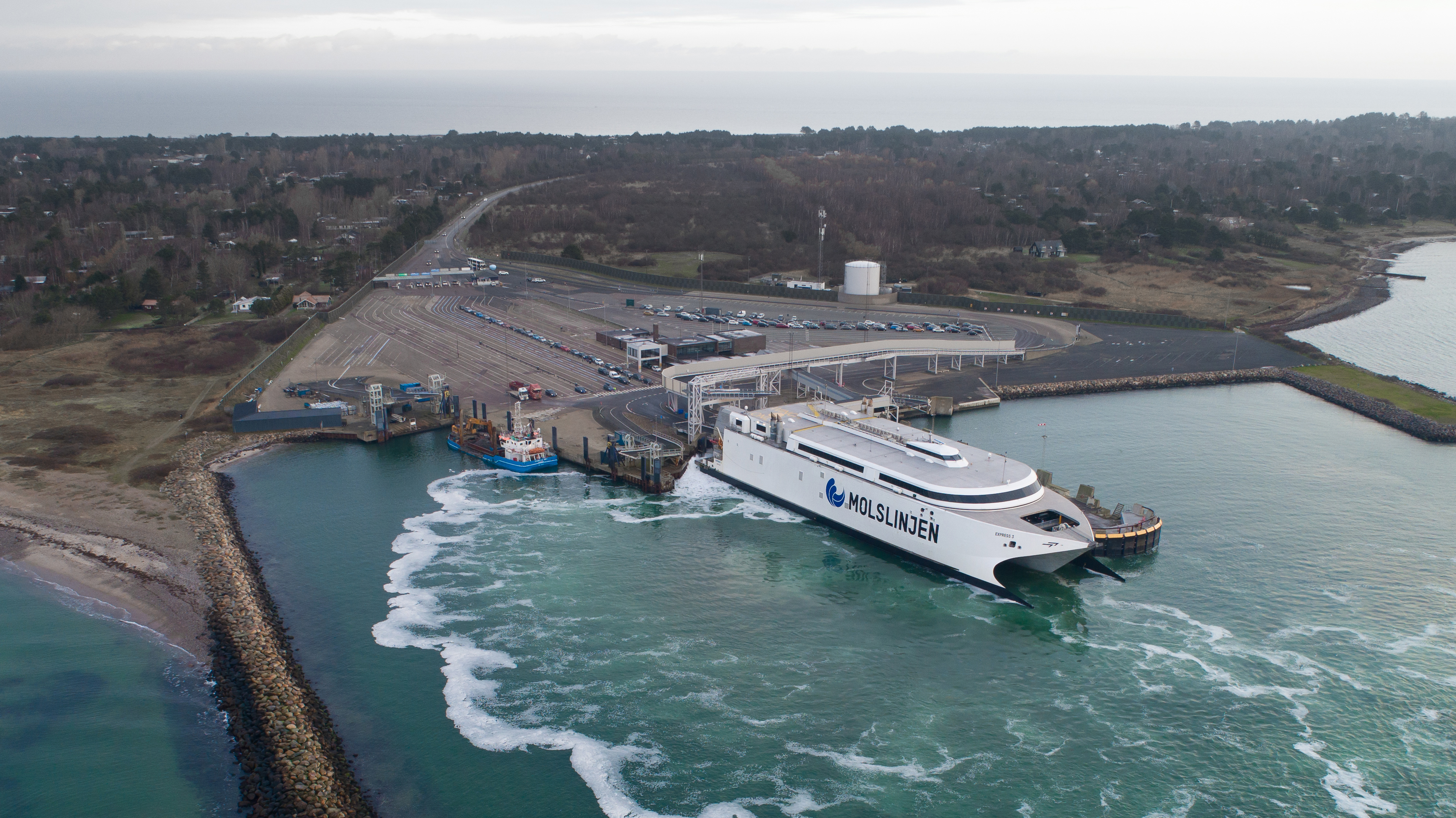 Molslinjen færge i havn ved kysten, set fra luften, omgivet af havvand og mole, klar til afgang.
