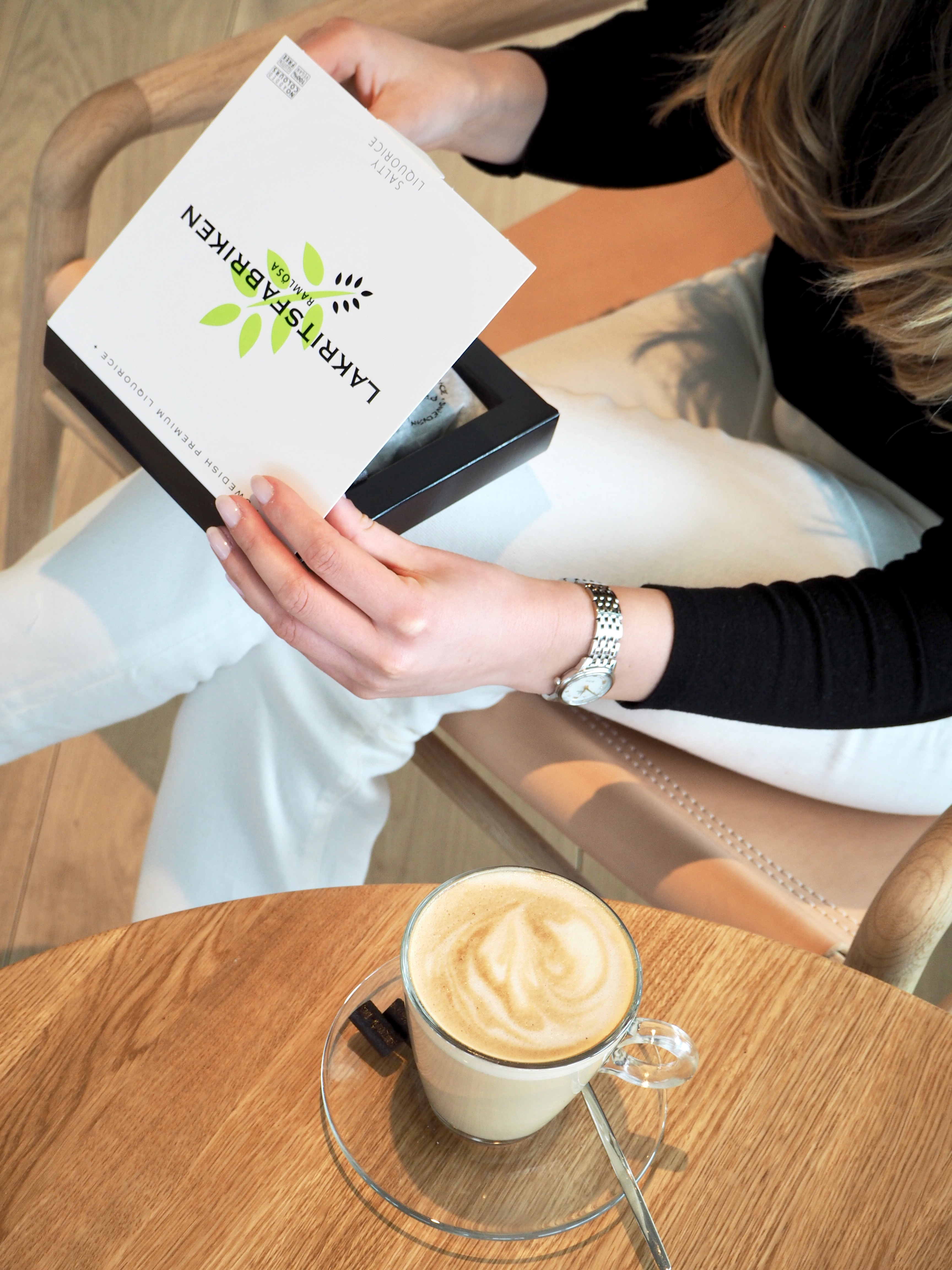 En person åbner en æske med lakridskonfekt fra Lakrids by Bülow ved et cafébord med cappuccino.