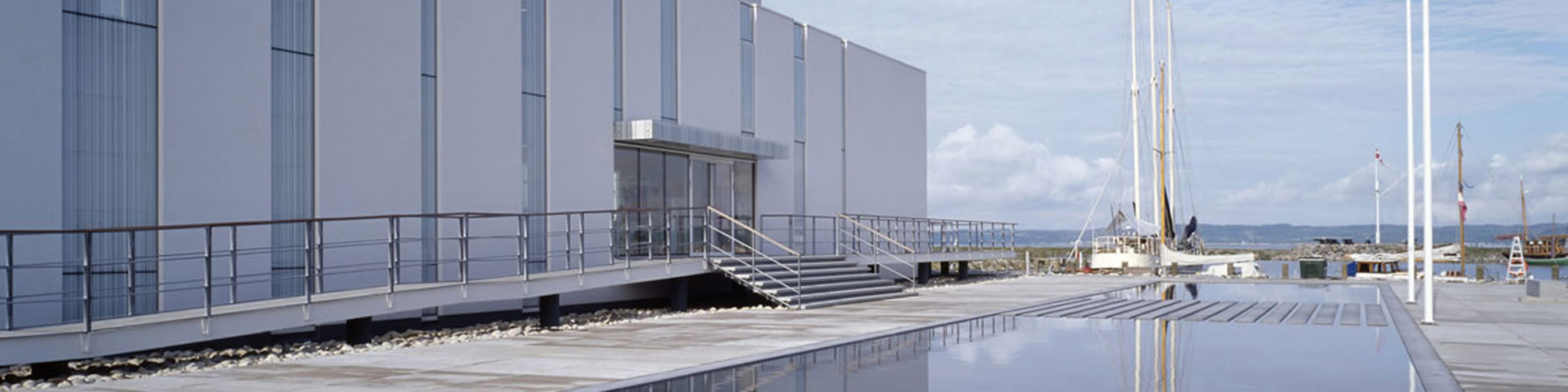 Moderne bygning med glasindgang og stålhåndlister ved havnefronten under en klar blå himmel.