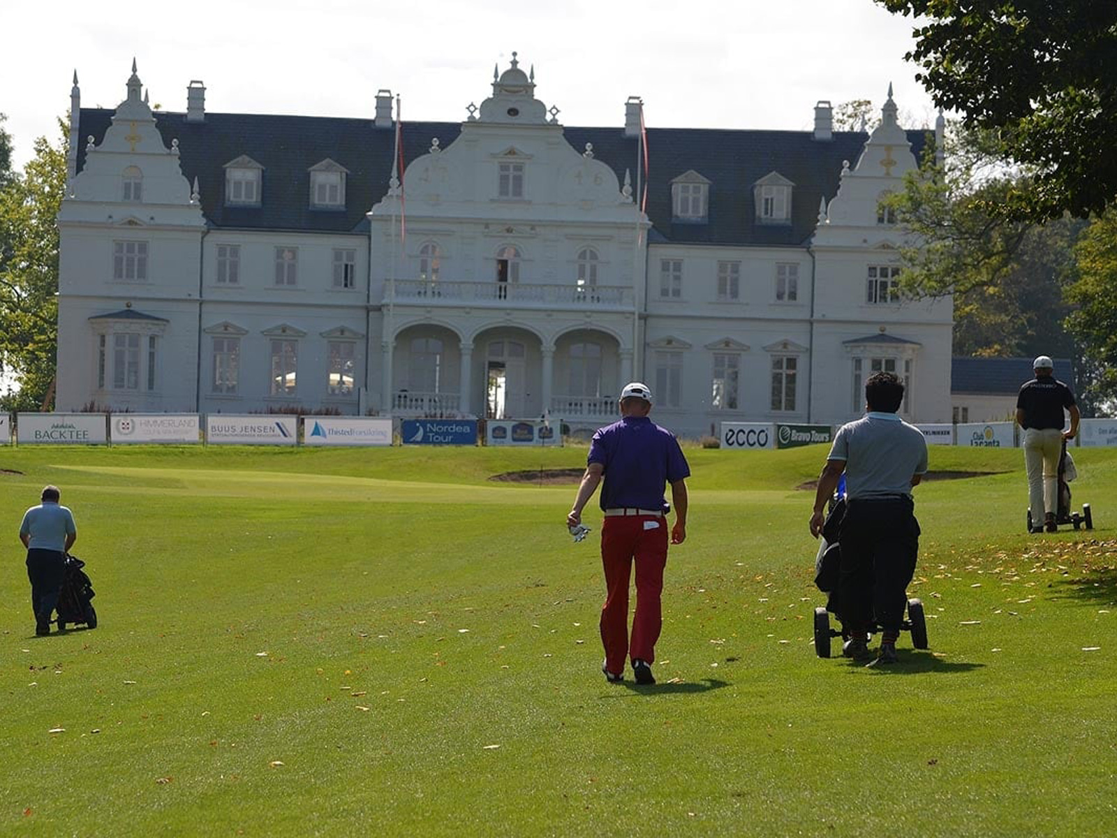 Golfspelare på en grön fairway i förgrunden av Kokkedal Slotts majestätiska fasad en solig dag.