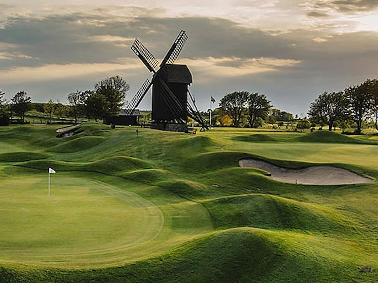 Golfbane med vindmølle i baggrunden under overskyet himmel, grønne fairways og sandbunker i forgrunden.