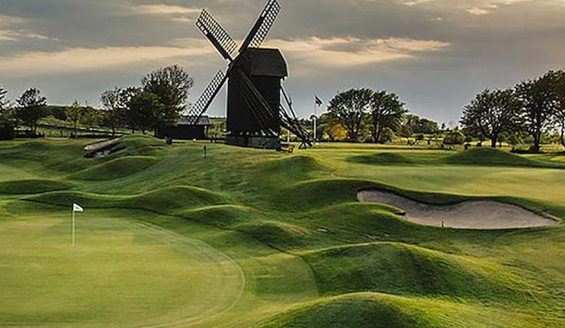 Golfbane med vindmølle i baggrunden under overskyet himmel, grønne fairways og sandbunker i forgrunden.