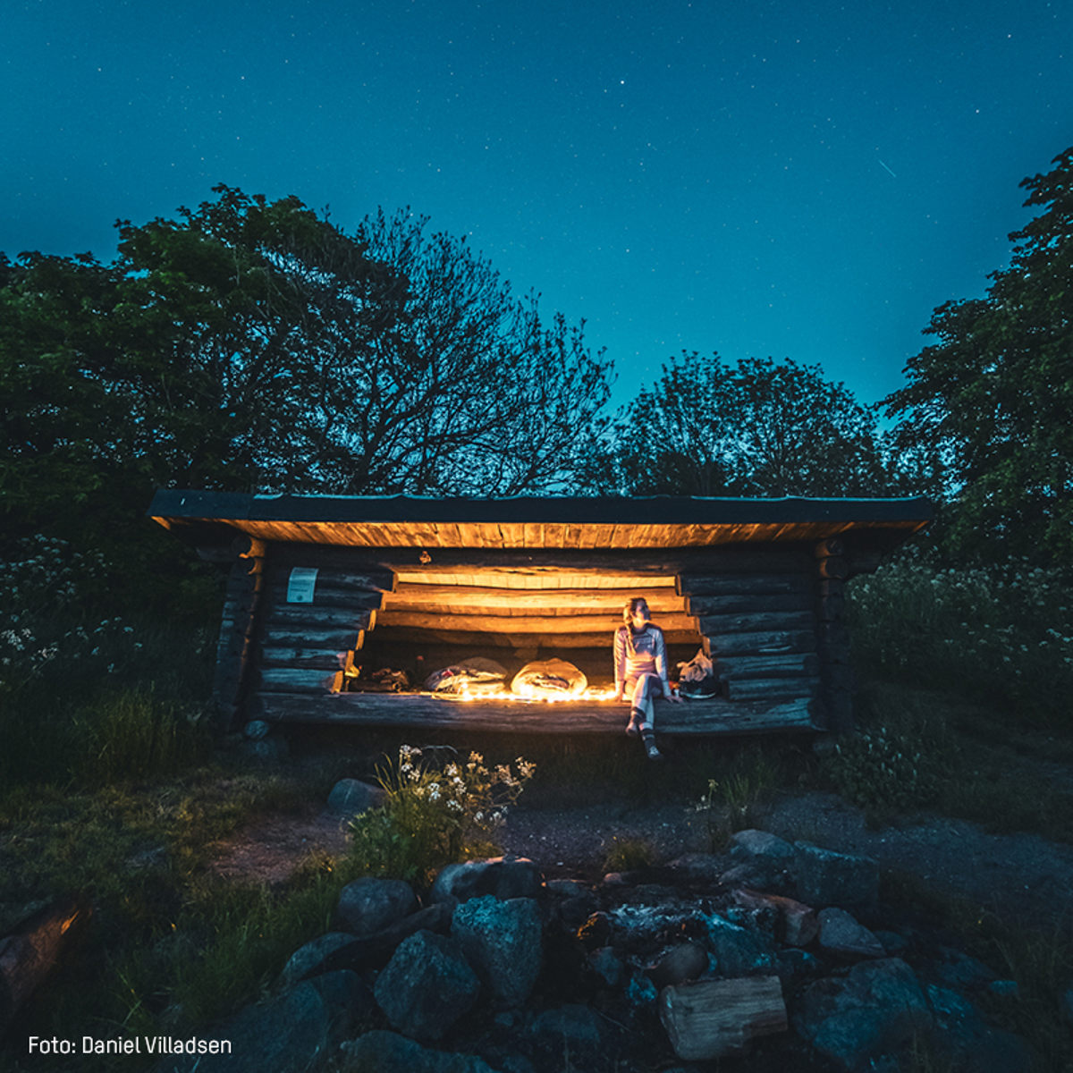 Person sidder foran bål i en træhyggeshelter om natten, omgivet af skov.