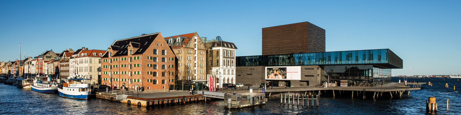 Københavns Skuespilhus ved havnefronten i solrigt vejr med udsigt over vandet og historiske bygninger.