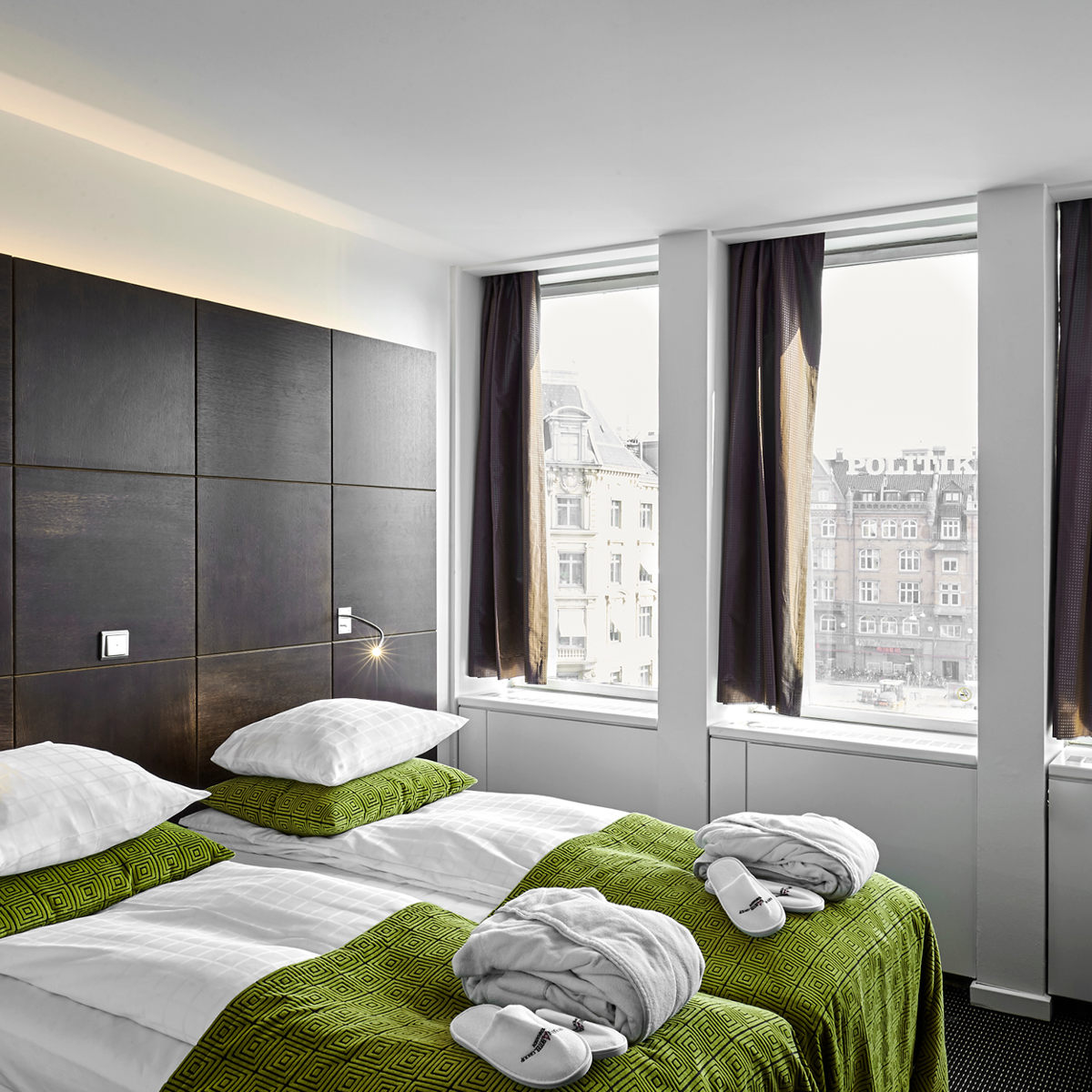 To senge med grønne sengetæpper i et moderne hotelværelse med store vinduer og udsigt til byen.