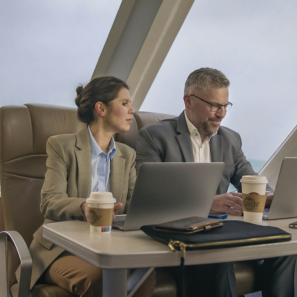 To personer arbejder på bærbare computere ved et bord på en færge med kaffekopper i hånden.