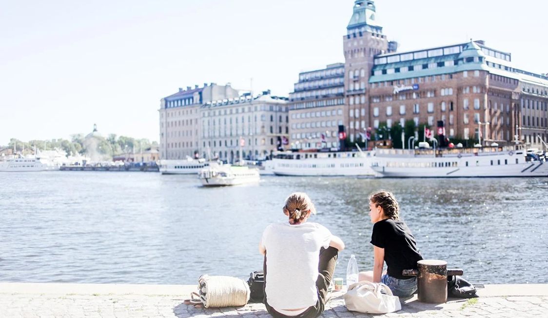 To personer sidder ved vandet med udsigt til ældre bygninger og både, solskin skaber en rolig atmosfære.