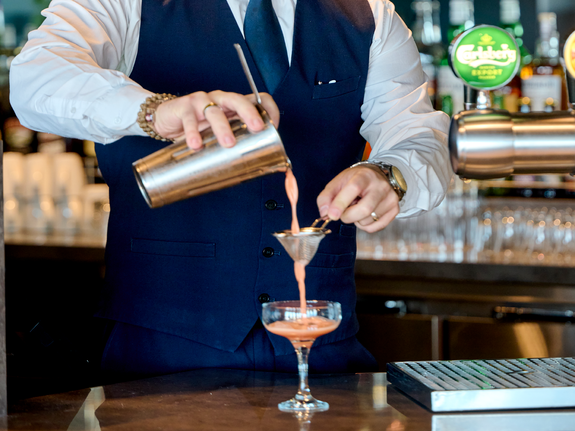Bartender i blå väst häller rosa cocktail från shaker genom sil i glas på bar.