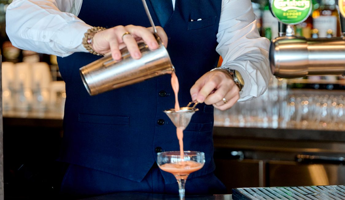 Bartender i blå väst häller rosa cocktail från shaker genom sil i glas på bar.