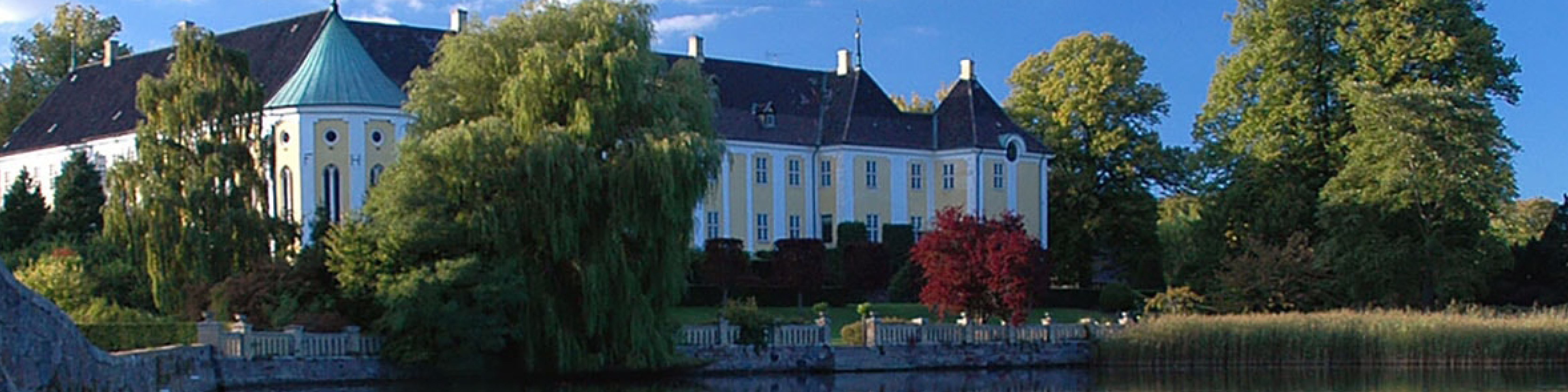 Gult historisk slot med hvidt tårn og skovset i baggrunden ved søbred i solskin.