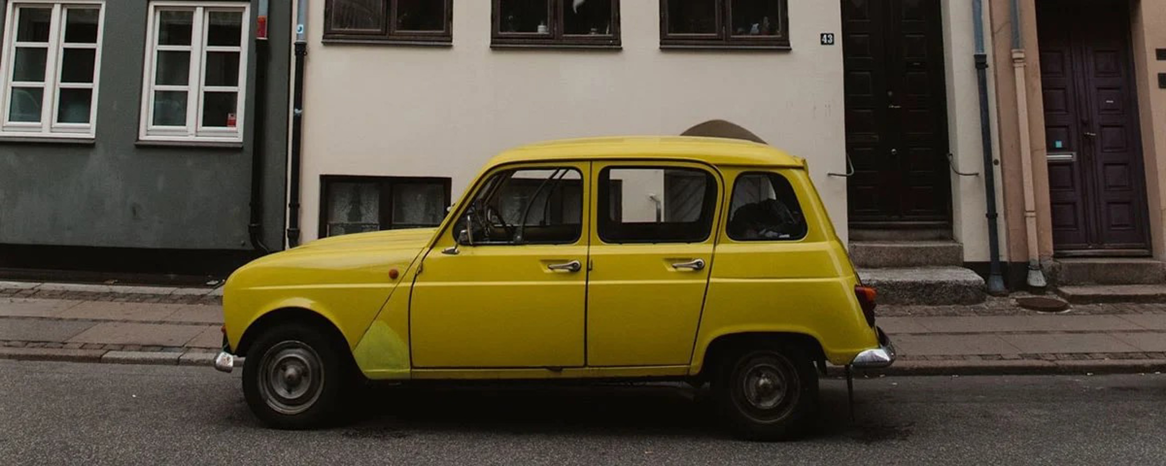 Gul retrobil parkerad på gata framför byggnad med vita och mörka fönster i urbana omgivningar.