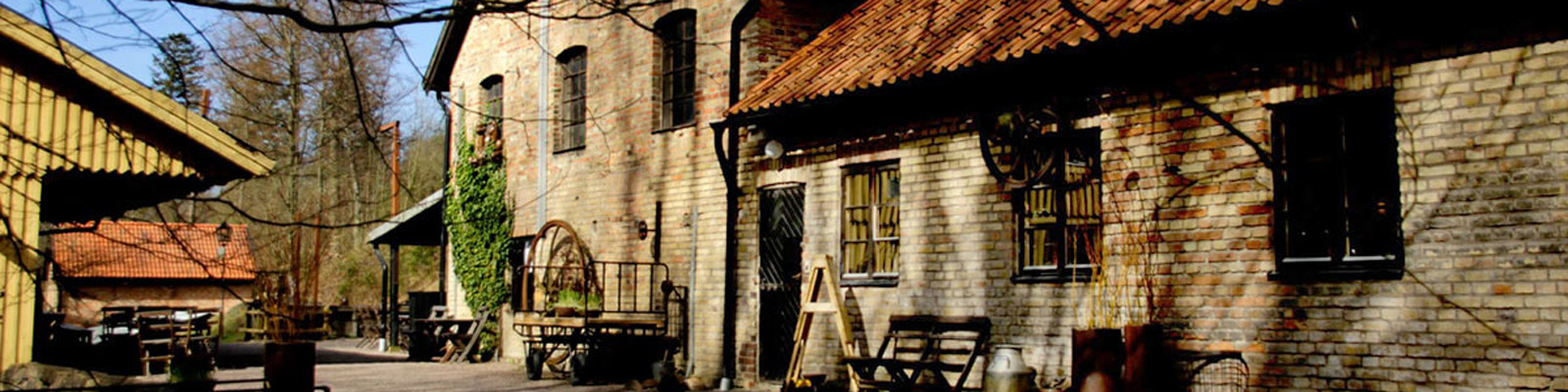 Gammelt dansk murstenshus med rødt tegltag og trærammer beliggende i skovklædt område under klar himmel.