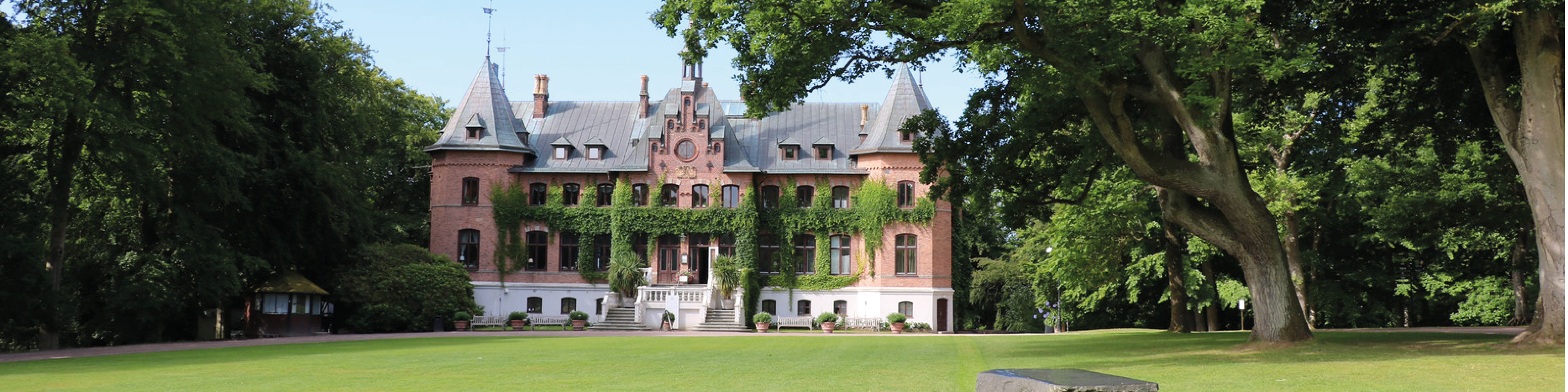 Stort slot i klassisk arkitektonisk stil beliggende i frodig have omkranset af træer i sommermånederne.