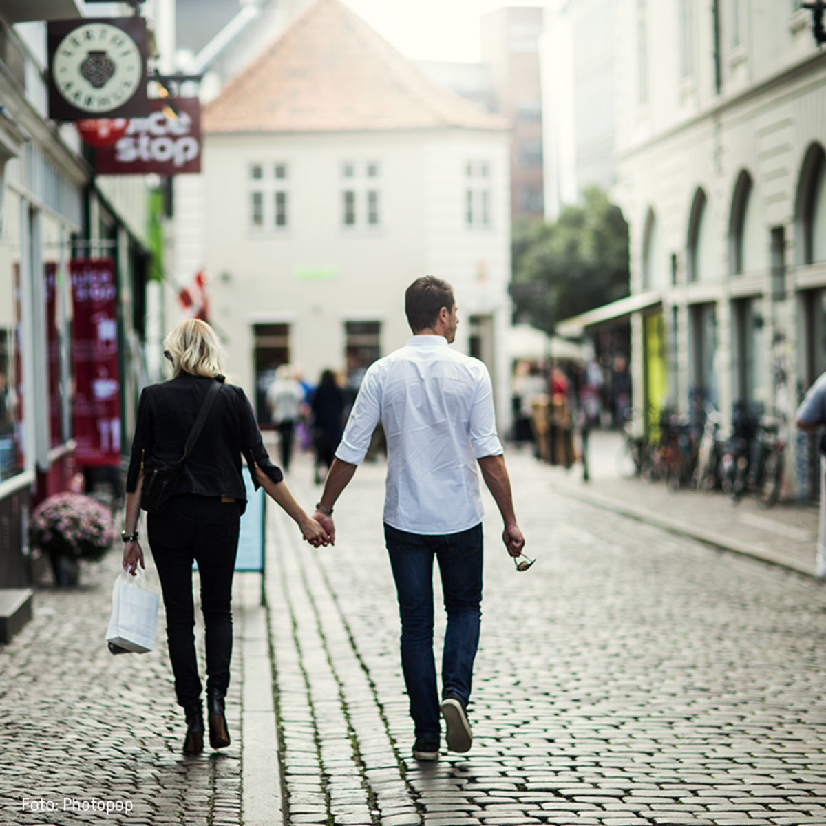 Et par går hånd i hånd på en brostensbelagt gade i en by, omgivet af butikker og cykler.