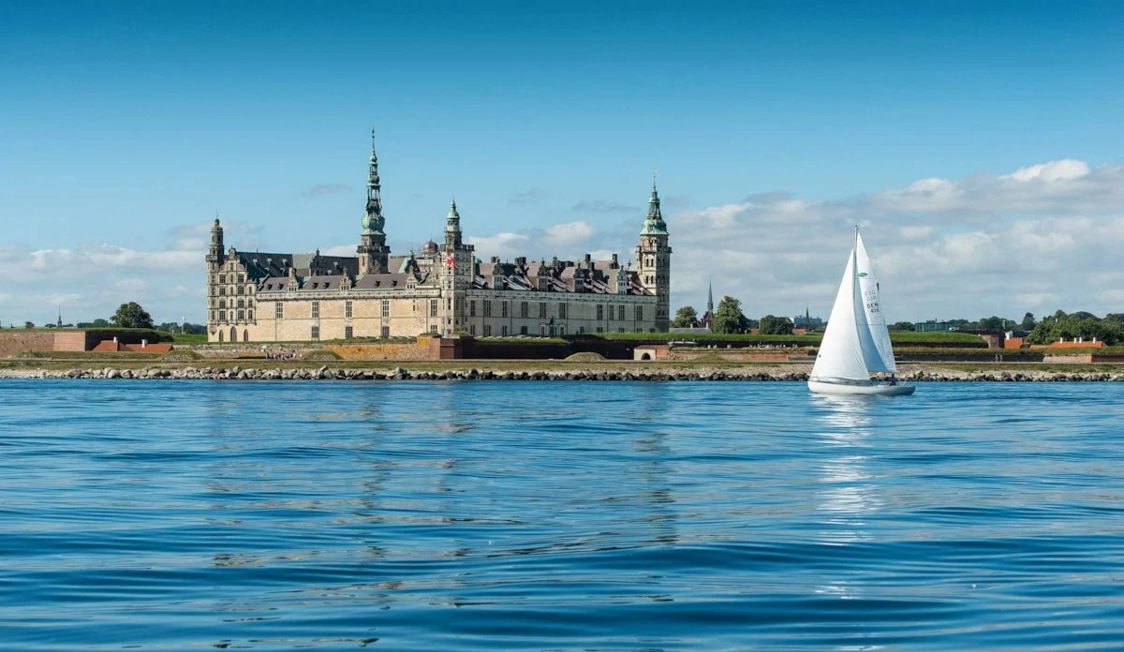 Vit segelbåt seglar förbi Kronborgs slott med lugna vatten och klarblå himmel i bakgrunden.
