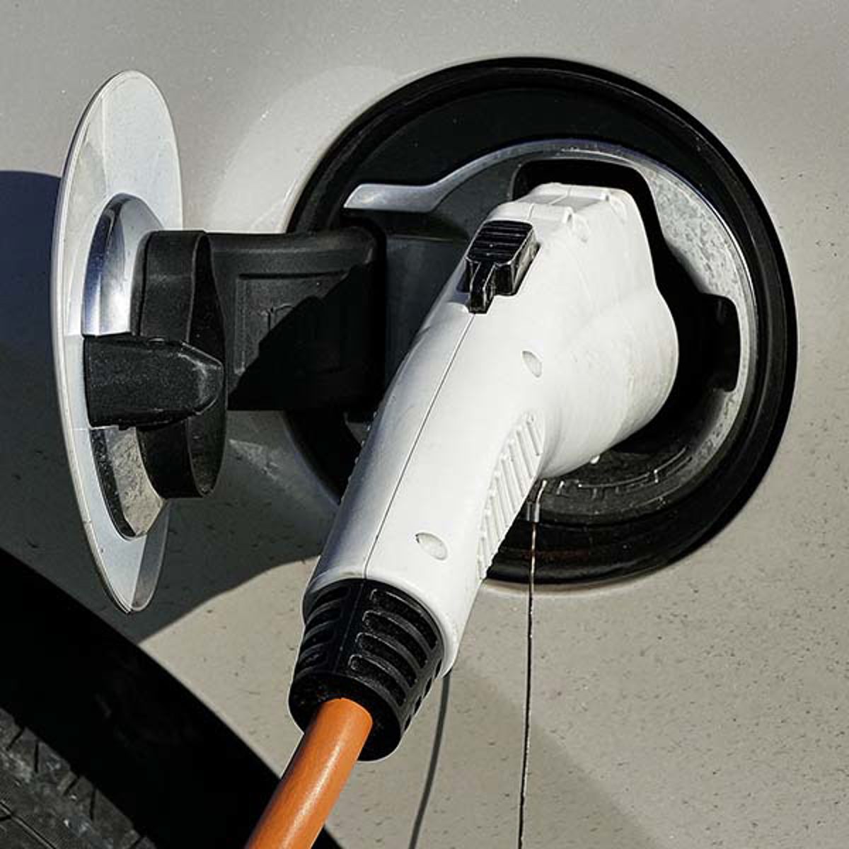 Hvid elbil oplader tilsluttet til køretøjens ladestik med orange kabel foran sølvfarvet bil.