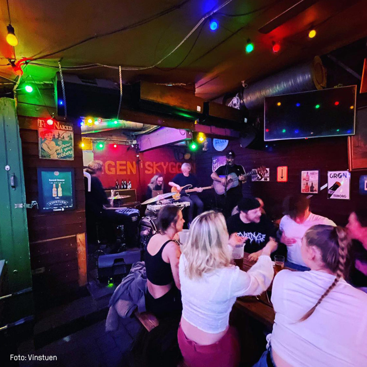 Live band spiller i hyggelig bar, farverige lys og glade gæster nyder atmosfæren ved borde.