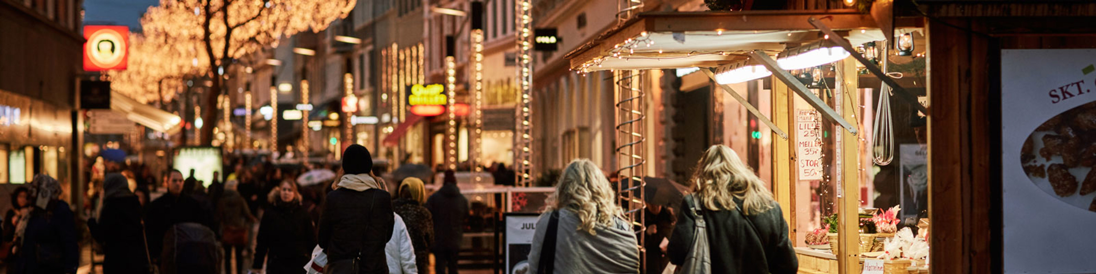 Shoppinggade oplyst med julebelysning og mennesker, der går forbi en trækiosk om aftenen.