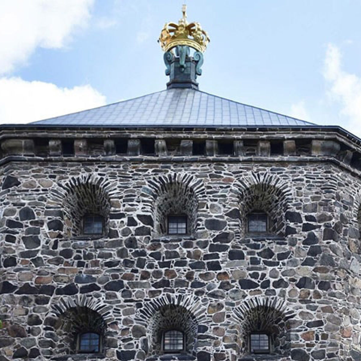 Stenbygning med gyldent tårn og skiffer tag under blå himmel.