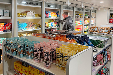 Butikshylder fyldt med forskellige snacks og slik, mens en kunde i grå jakke rækker ud efter en pose slik.