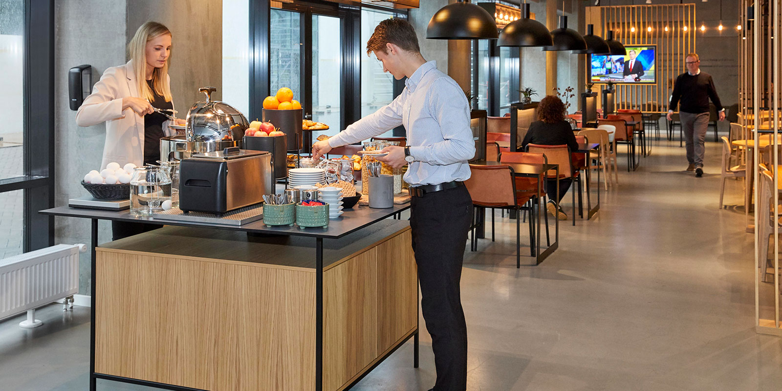 Mand klædt i skjorte fylder tallerken ved morgenmadsbuffet med frisk frugt og bagværk i moderne spisestue.