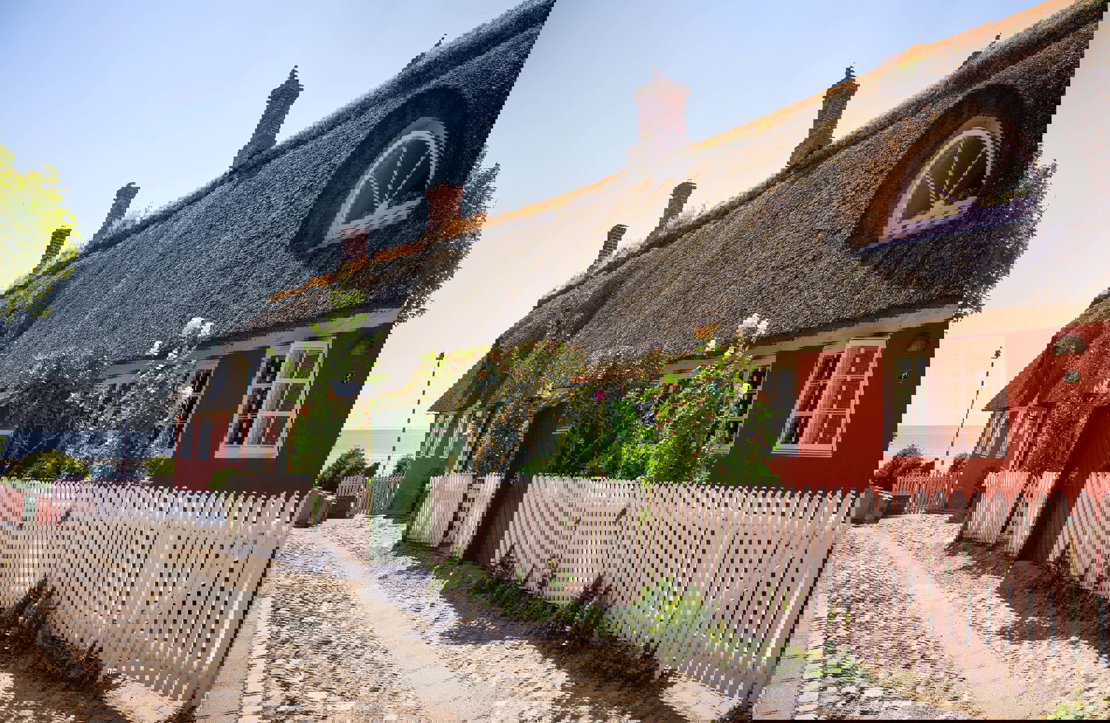 Rødt hus med stråtækttag ses med hvid stakit i forgrunden.