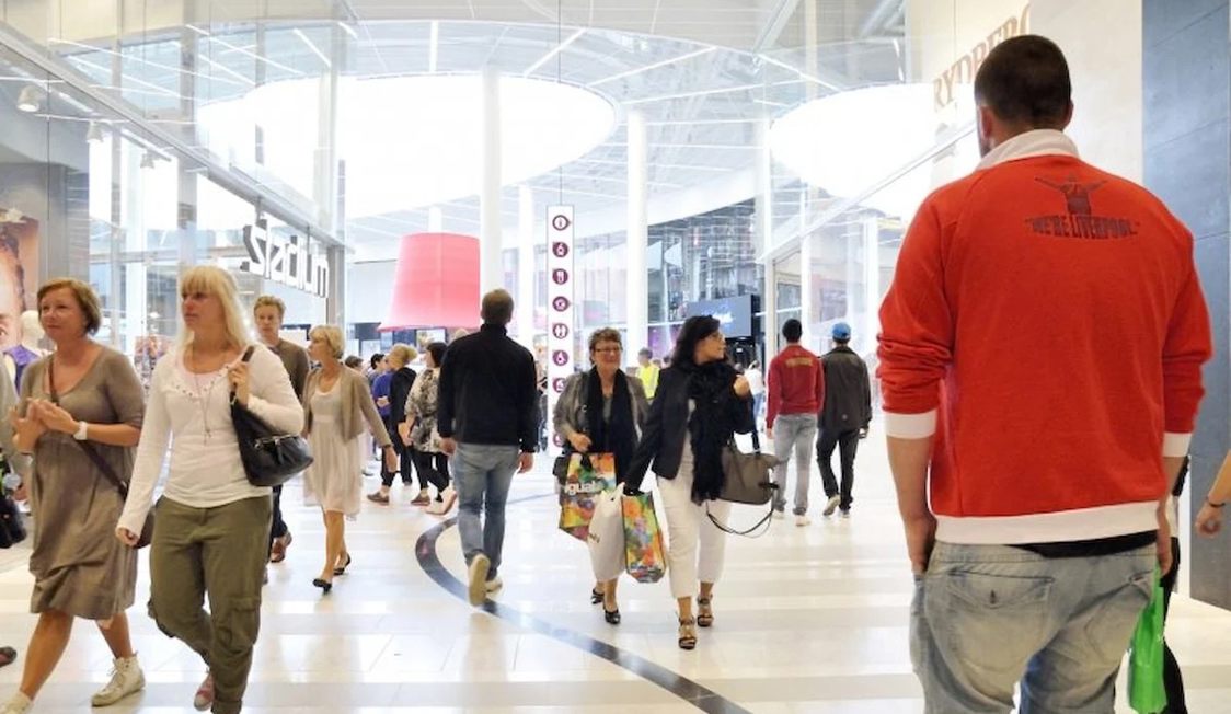 Folkemængde går gennem indkøbscenter med glasloft, forskellige butikker og tydelig skiltning i baggrunden.