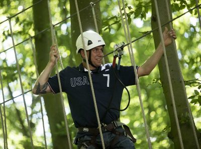 Mand klatrer i højder på en rebparkbane iført sikkerhedsudstyr og hjelm i en skovrig omgivelser.
