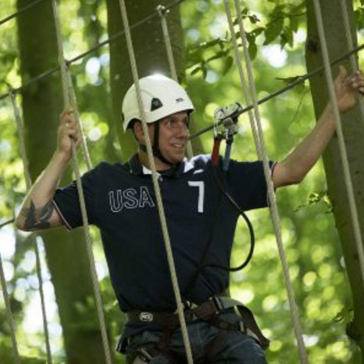 Mand klatrer i højder på en rebparkbane iført sikkerhedsudstyr og hjelm i en skovrig omgivelser.