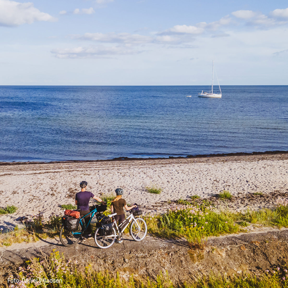 To cyklister med hjelme nyder udsigten over havet fra en sti nær en sandstrand i sollys.