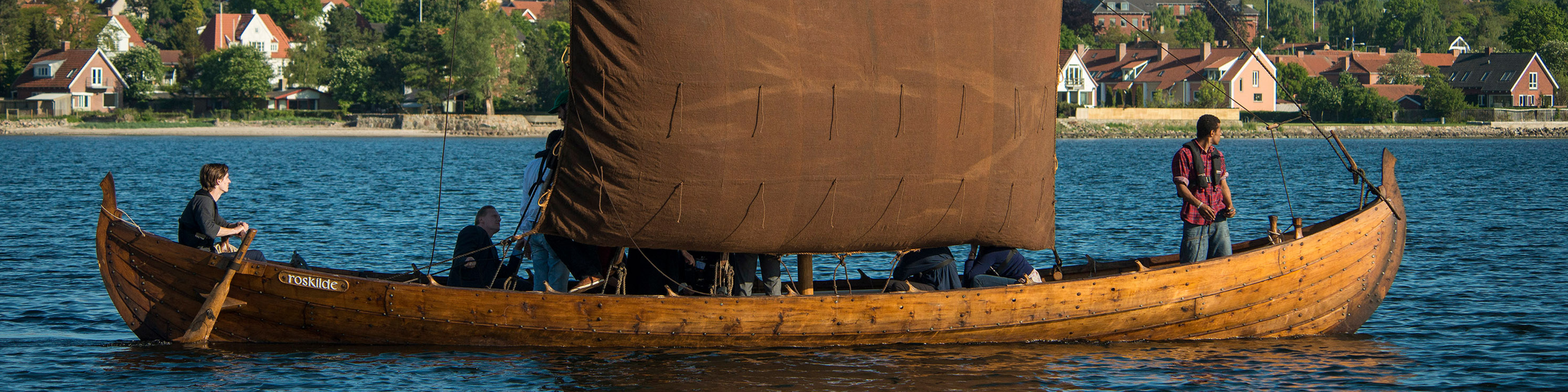 Trævikingeskib med brun sejl sejler på fjorden med besætning ombord, set fra siden med by i baggrunden.