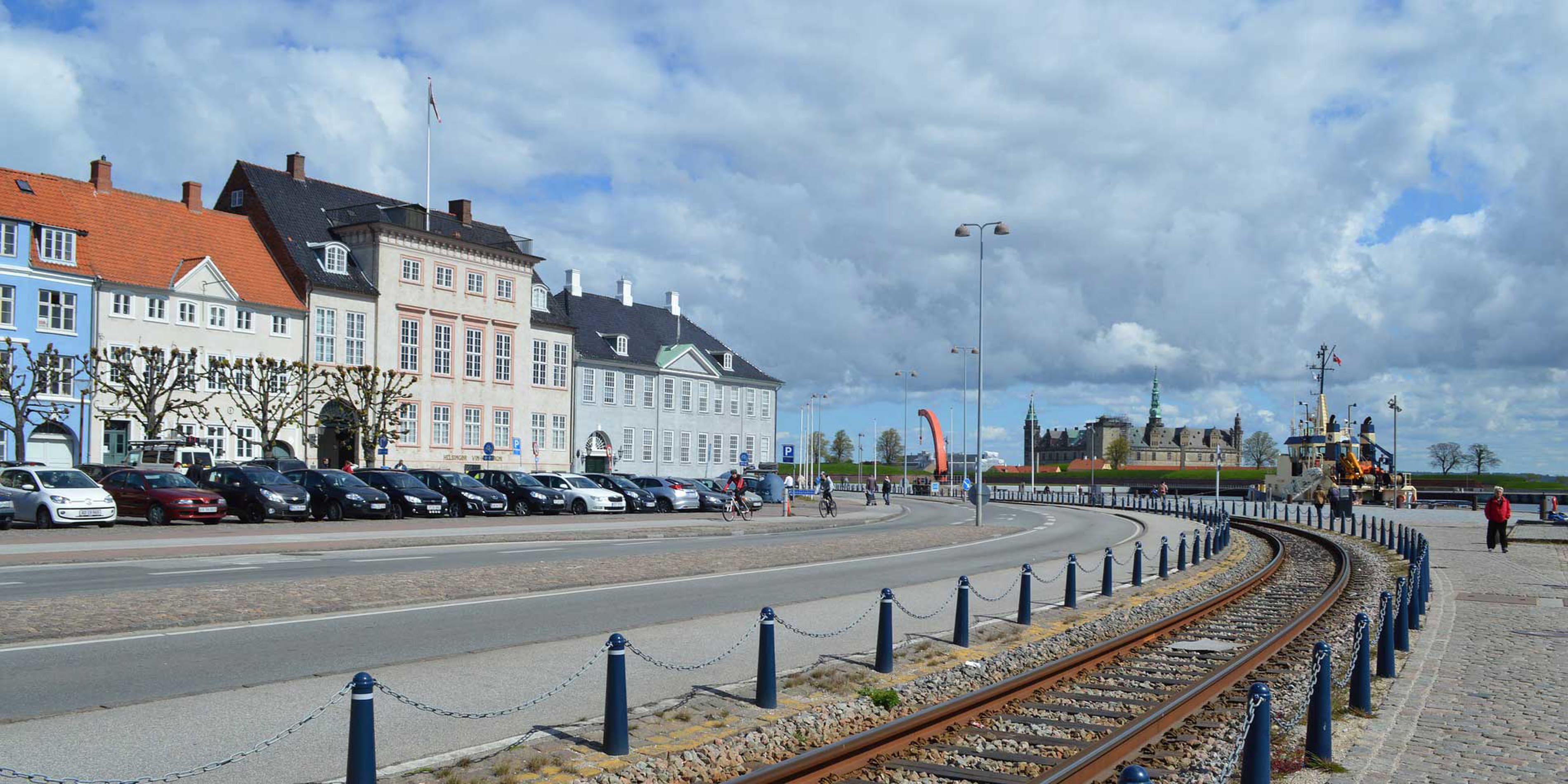 Bilar parkerade framför färgglada byggnader på en gata med spår mot Kronborgs slott på en molnig dag.