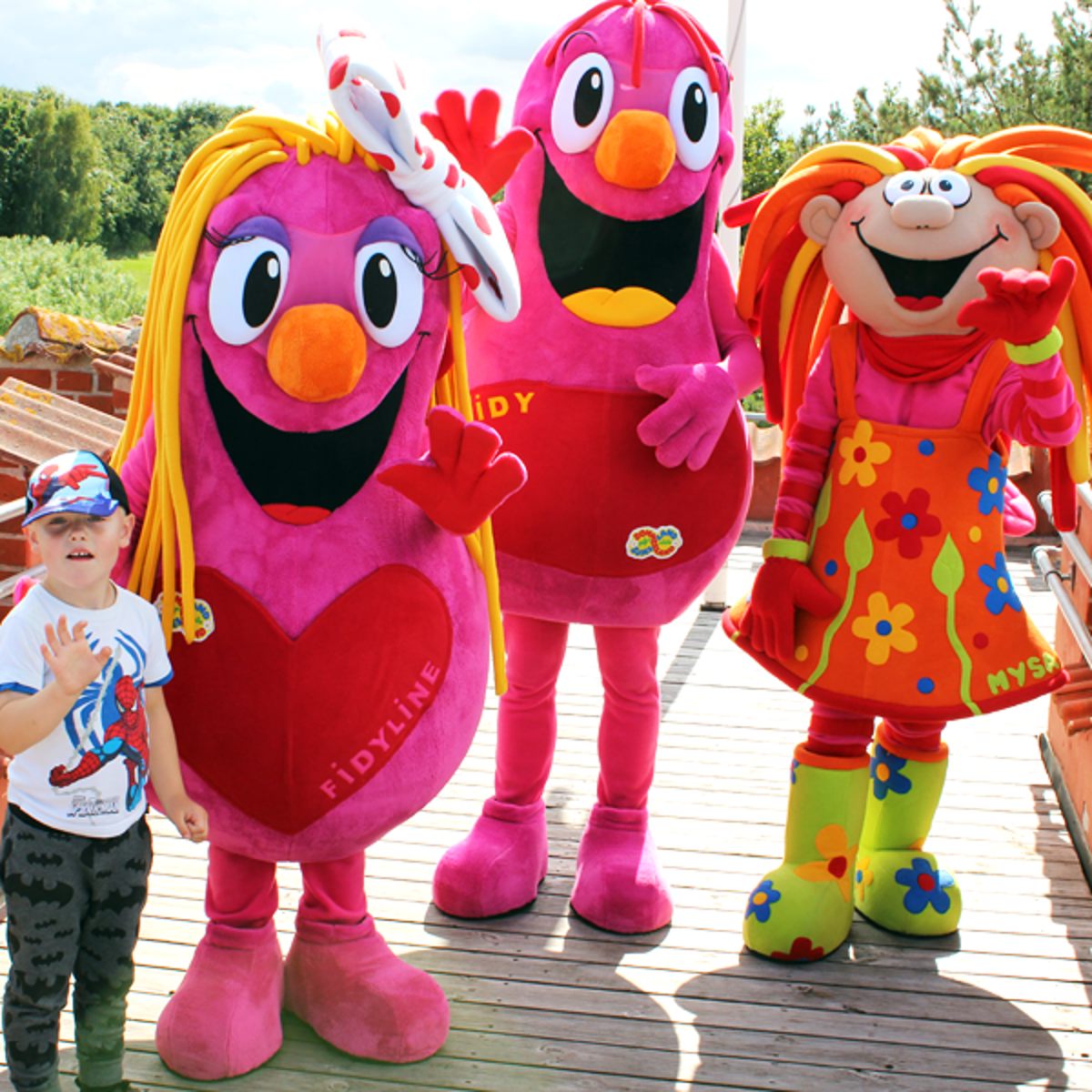 Tre farverige maskotter i sjove kostumer hilser glade på et barn udenfor på en solrig dag.