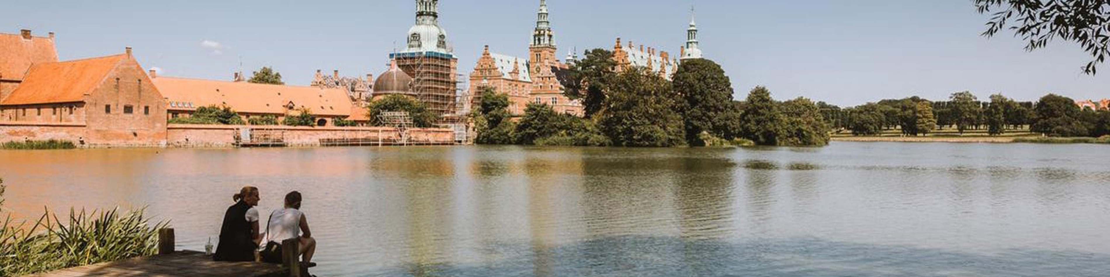 Frederiksborgs slott vid sjö med två personer på brygga i Hillerød, sommardag i Danmark.