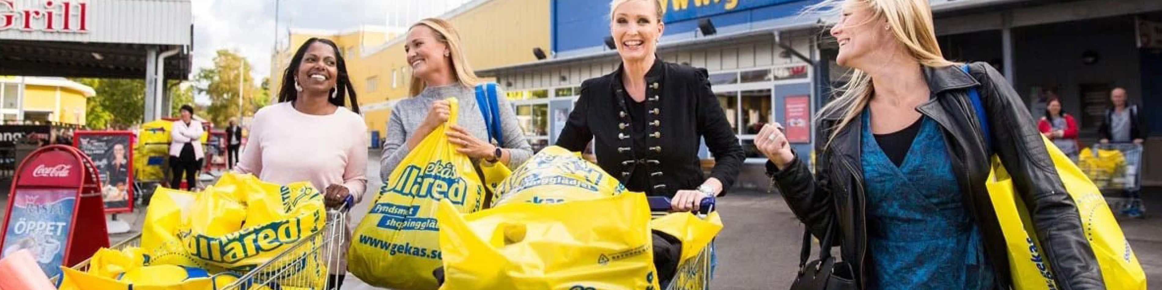 Fire glade kvinder shopper med store, gule poser uden for Gekås Ullared-butikken på en solrig dag.