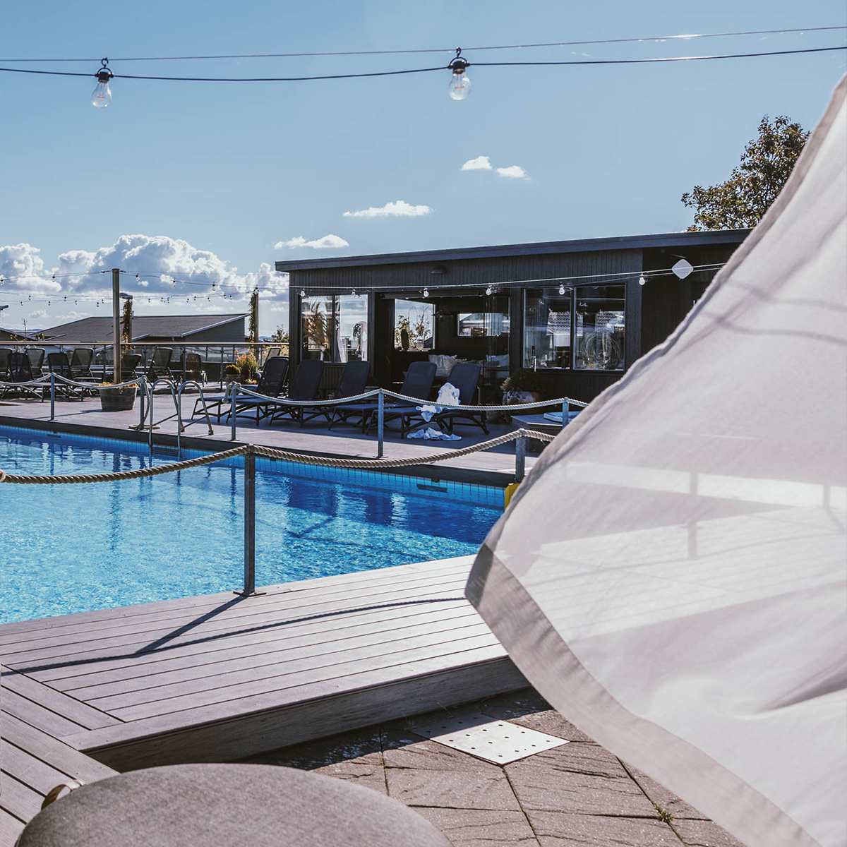 Udendørs swimmingpool ved moderne bygning med liggestole på træterrasse under blå himmel.
