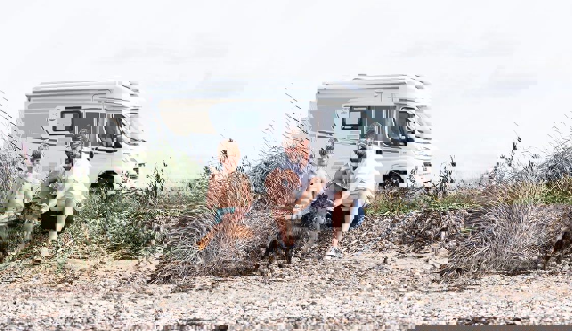 Två personer och en hund sitter på en strand med en skåpbil i bakgrunden under ett campingäventyr.