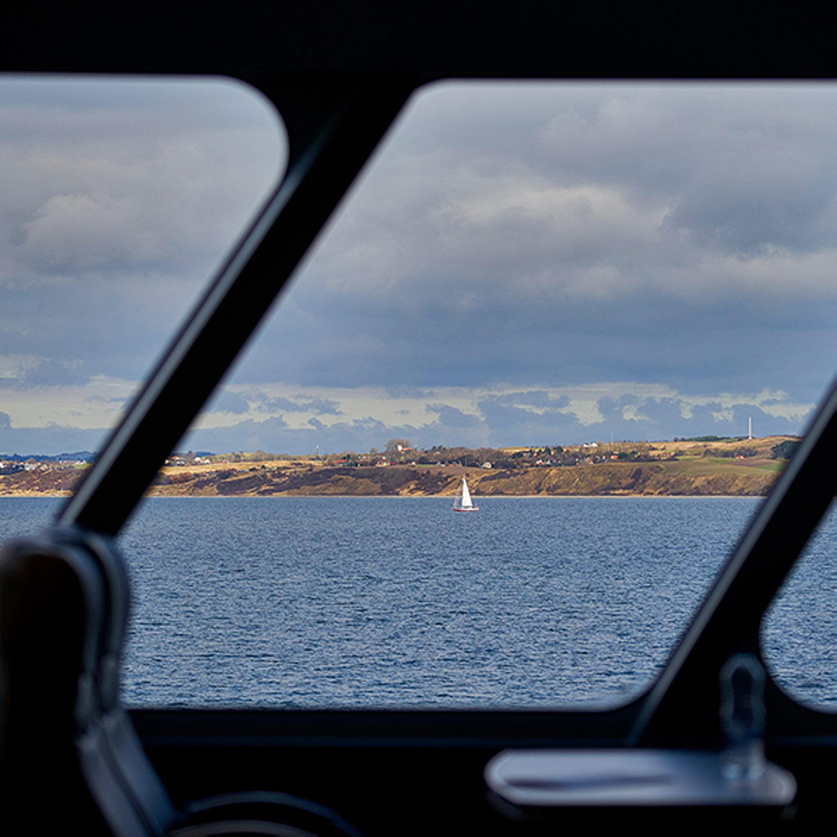 Sejlbåd på havet set fra færgens vindue, med kystlandskab i baggrunden under overskyet himmel.