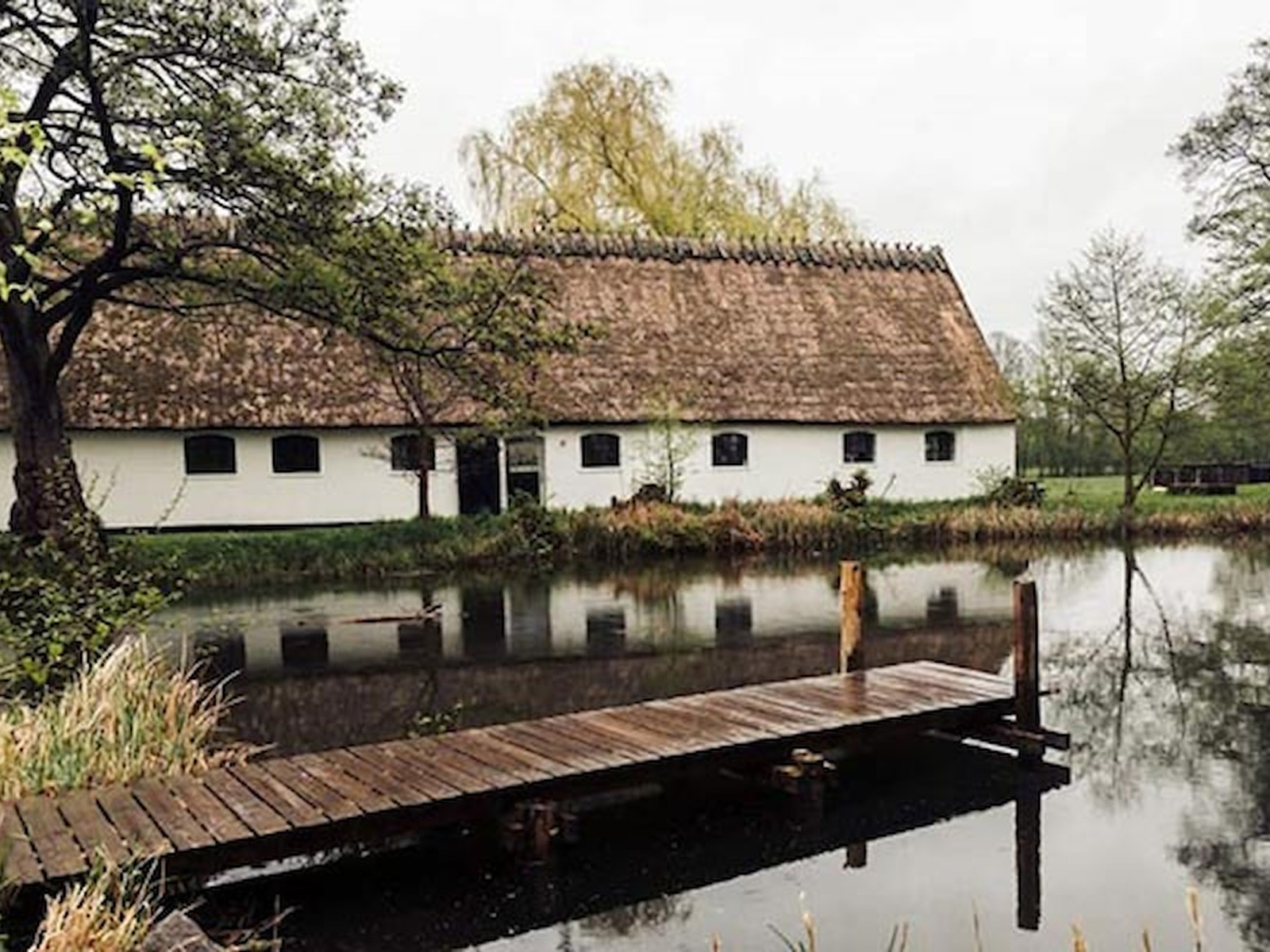 Esrum Mölgård brygga med vass tak längs en lugn damm omgiven av grönska och träd.