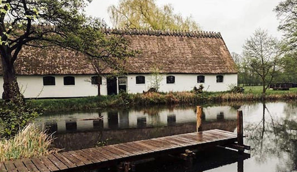 Esrum Mölgård brygga med vass tak längs en lugn damm omgiven av grönska och träd.